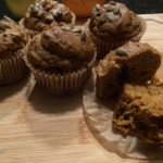 Freshly baked cranberry pumpkin muffins topped with pumpkin seeds on a cooling rack, surrounded by fall leaves and a small bowl of maple cream cheese.