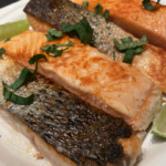 Crispy pan-seared fish filet topped with avocado sauce, diced tomato, and cilantro, served with a lemon wedge on a white plate.