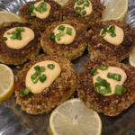 Crispy salmon cakes topped with Sriracha Aioli, garnished with green onions and lemon wedges on a white plate.